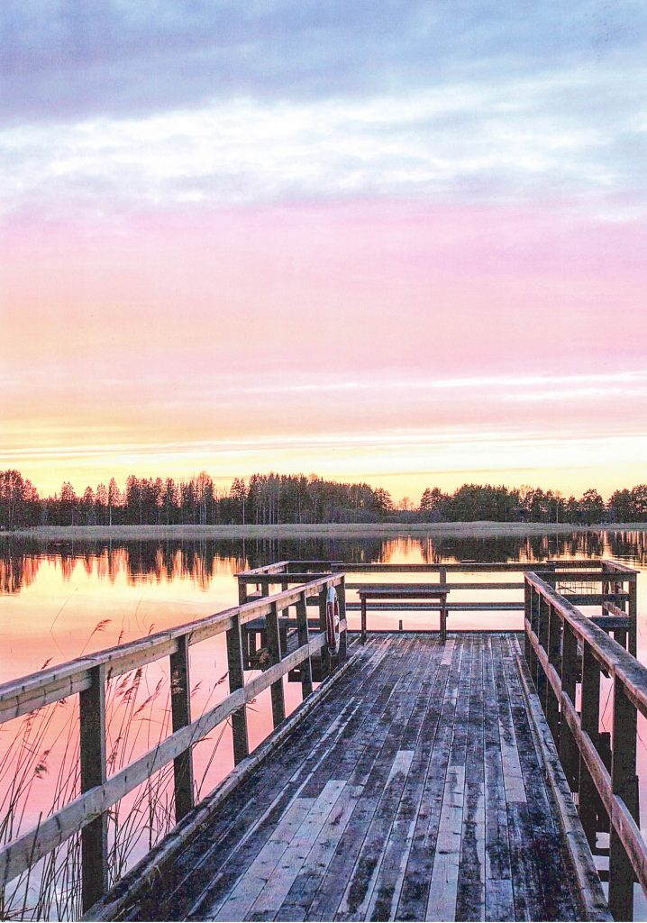 En träbrygga sträcker sig ut över stilla vatten i rosa och gyllene kvällsljus